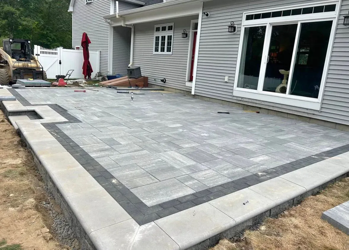Large gray stone patio with dark border beside a house, featuring sliding glass doors and a lawn edge
