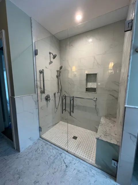 Accessible, marble-tiled shower with grab bars, built-in seat, and glass doors.