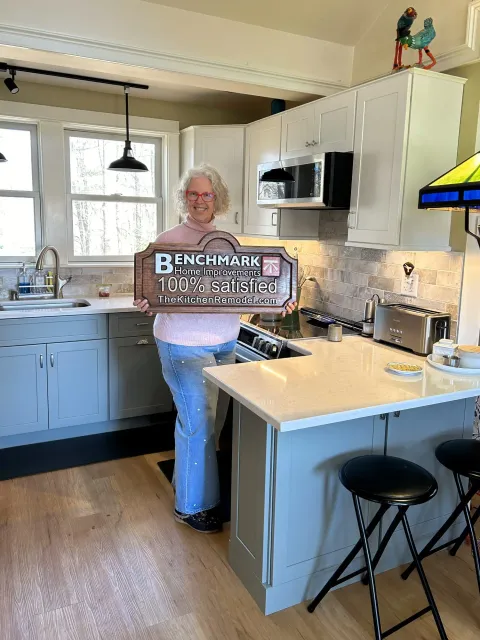 A homeowner holding a wooden Benchmark “100% Satisfied” sign in her finished sage-green kitchen