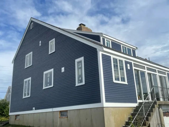 A blue two-story house with white trim is shown. The house features multiple windows, both on the main and upper floors. A staircase with a metal railing leads to the entrance on the right side. The roofing and siding complement each other beautifully against the partly cloudy sky.