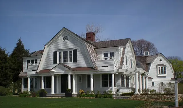 Large white farmhouse with a front porch, black shutters, and a green lawn in daylight