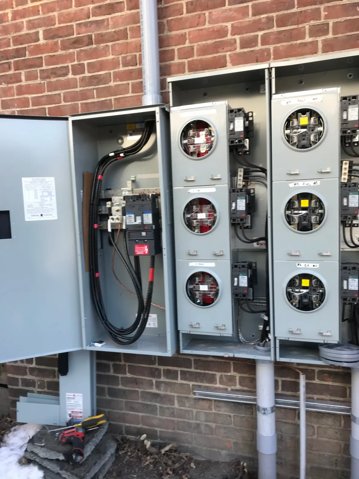 Electrical panels with open and closed circuit breakers and wiring, mounted on a brick wall, with tools on the ground nearby.