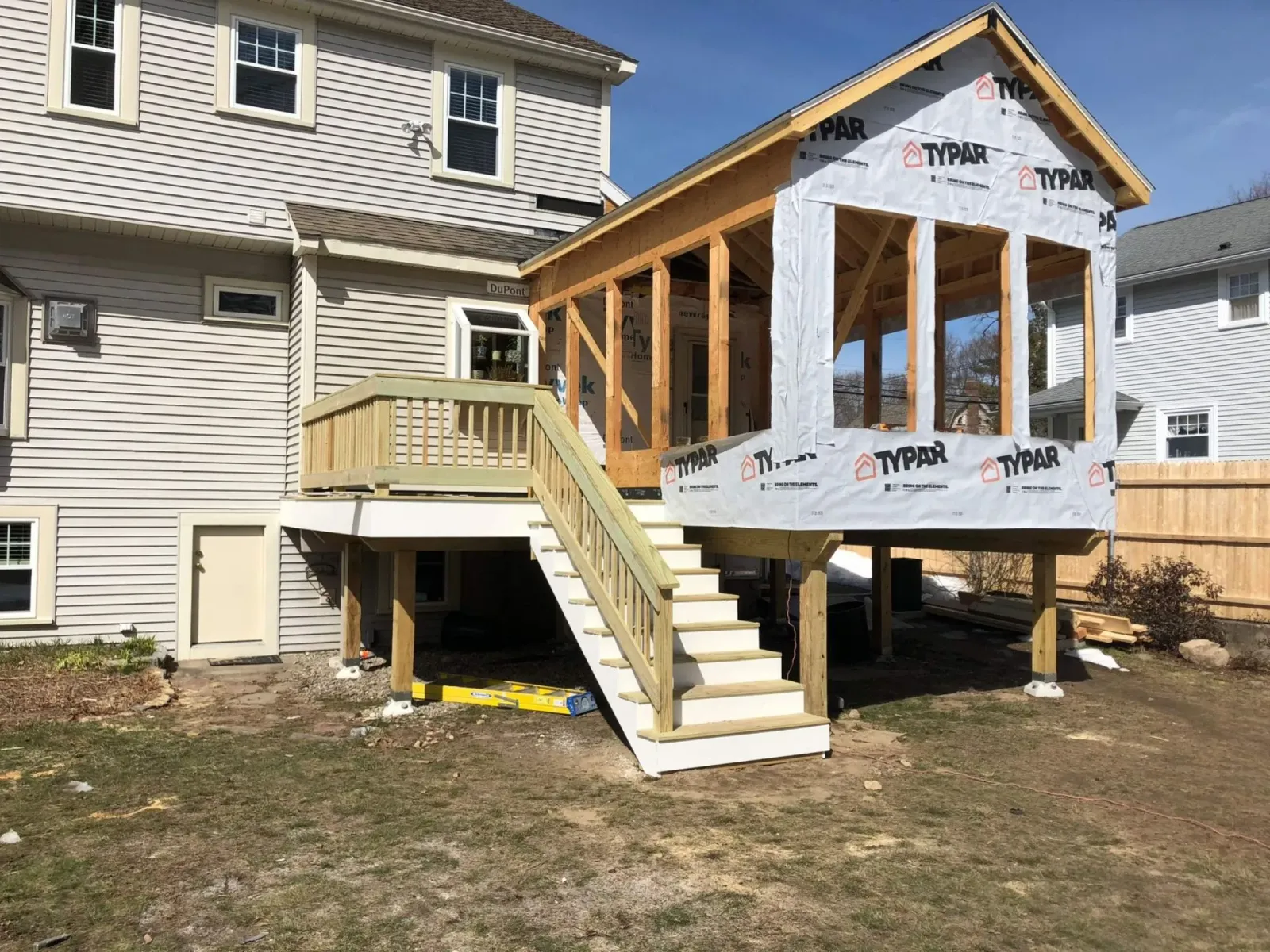 Wooden deck and sunroom addition to a light-colored house, under construction with Typar wrap, on a sunny day.