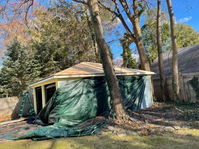 A small building covered in green tarp, likely for weather protection. Trees surround it in a yard.