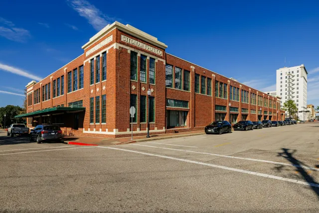 A large red brick building with green window frames, labeled 'STEDMAN FRUIT CO', under a clear blue sky. Multiple black cars are parked along the street in front of the building, with a tall white building in the background.