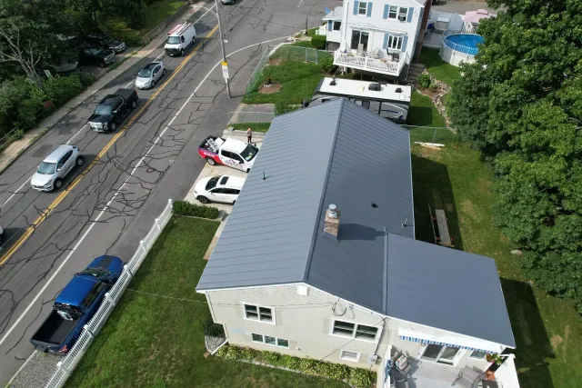 Aerial view of a house with a gray roof beside a street with parked cars and trees.