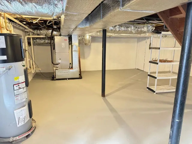 Empty basement with concrete floor, exposed pipes, water heaters, and a black support column.