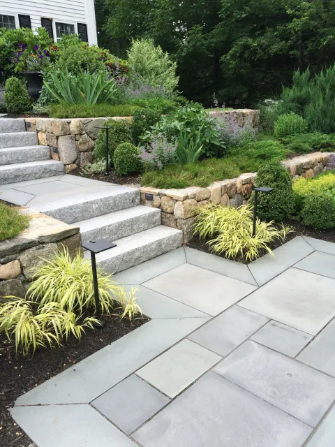 Granite steps and blue stone walkways combined with a series of field stone retaining walls soften a steep slope, create a functional front door entry and offer dramatic curb appeal. The gardens are designed for four season interest in this North And