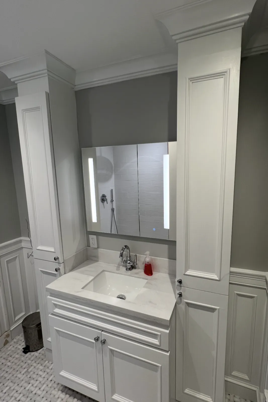 Bathroom vanity with a white countertop, a sink, and a large mirror with integrated lighting, surrounded by white cabinetry and gray walls. Installed by Total Home Services