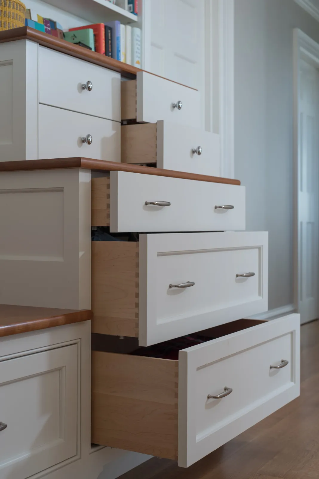 Open drawers of a white wooden dresser with silver handles in a room with bookshelf in the background.