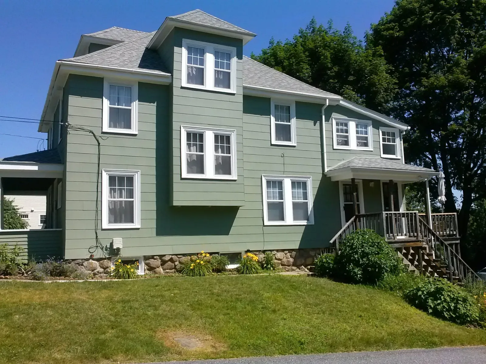 Green three-story house with white-framed windows, porch, and a stone foundation, set on a grassy lawn.