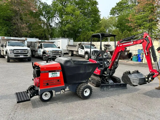 Red-and-black mini excavator with loader bucket in a gravel lot, trucks parked behind it