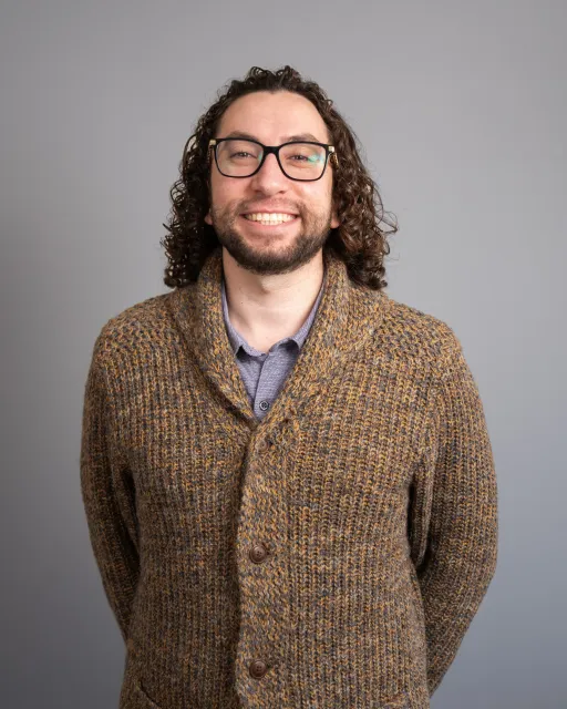 A man with curly hair, glasses, and a beard wearing a brown knitted cardigan smiling against a plain gray background.