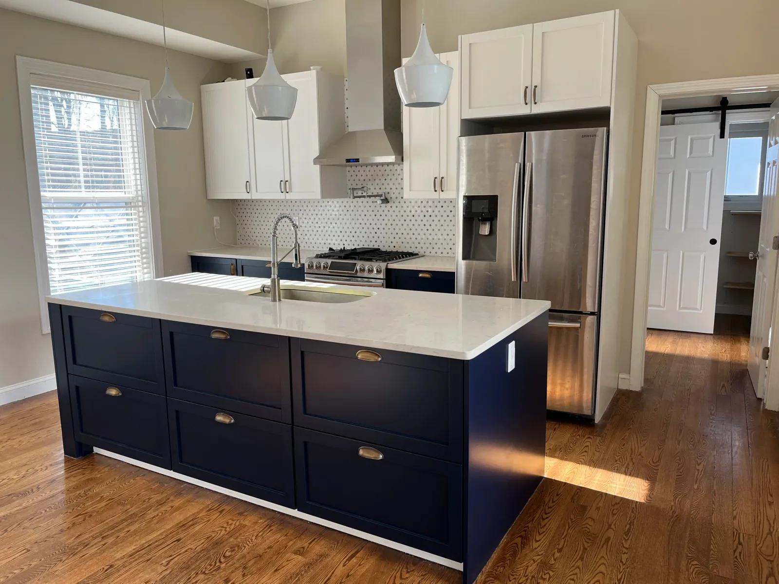 Modern kitchen with blue island, white cabinets, stainless steel refrigerator, oven hood, pendant lights, and wood flooring.