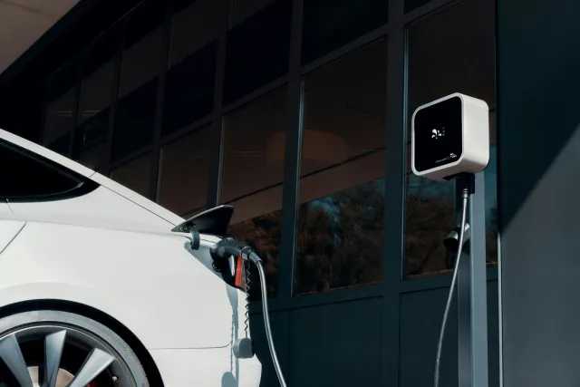 White electric car plugged into a charging station outside a modern building with large glass windows.
