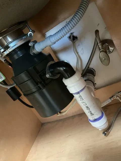 Under-sink view showing a garbage disposal unit connected to plumbing, drain pipes, flexible hoses, and a section of the sink drain pipe.