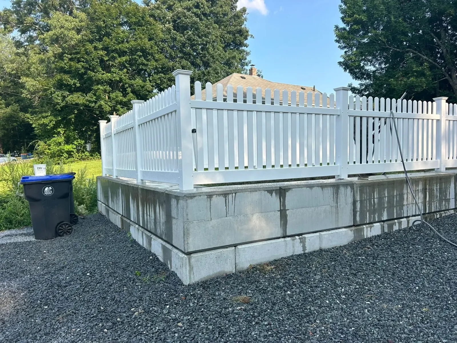 White fenced platform on a gravel lot beside trees and a blue trash bin