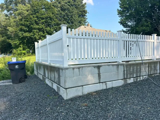 White fenced platform on a gravel lot beside trees and a blue trash bin