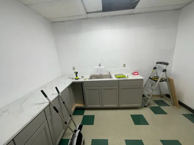 Empty kitchen with grey cabinets, marble countertop, stainless steel sink, and two ladders, one of which has a sign that says 'Out of Order'.