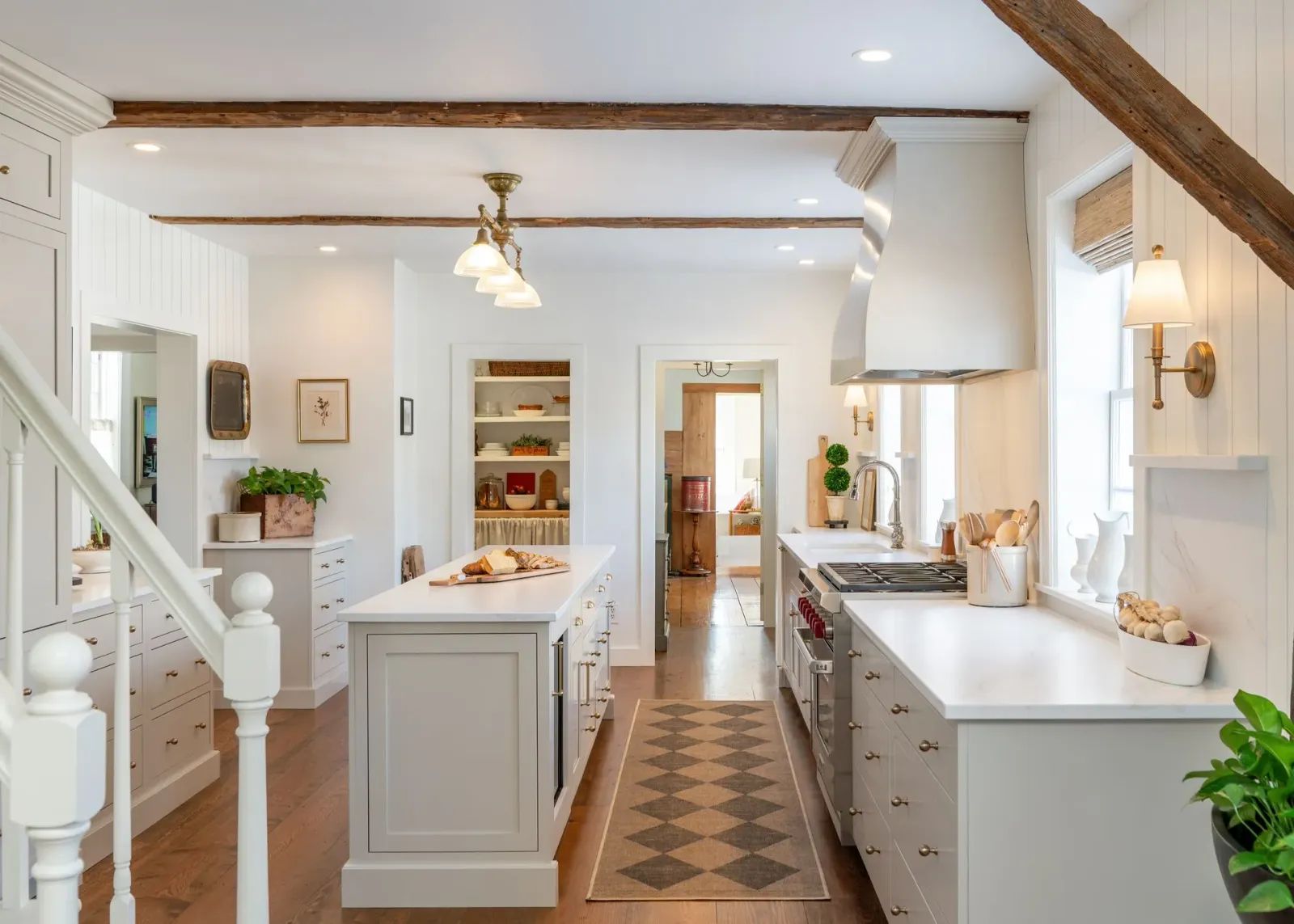 Bright white farmhouse kitchen with island, wood beams, patterned runner, and open doorway to a pantry.