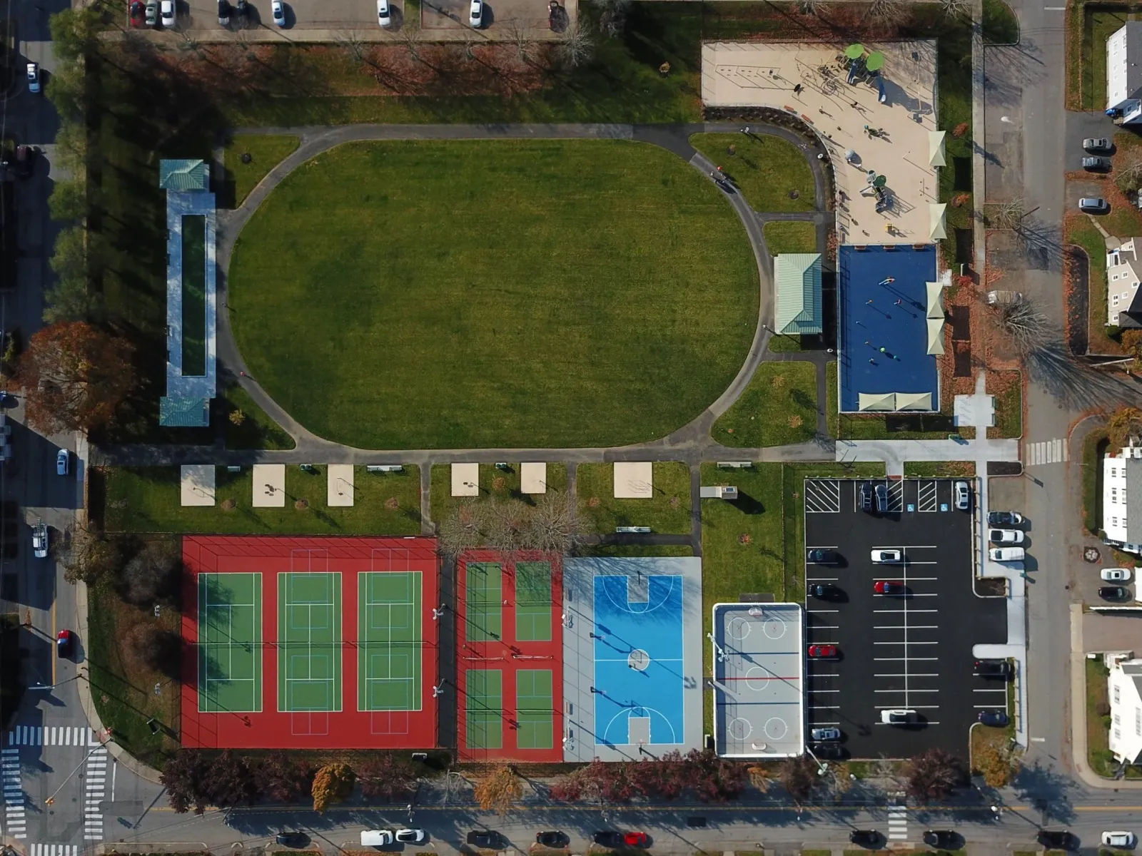 Aerial view of a community park featuring a large grassy oval field, multiple tennis courts, a basketball court, playgrounds, a parking lot, and surrounding streets.