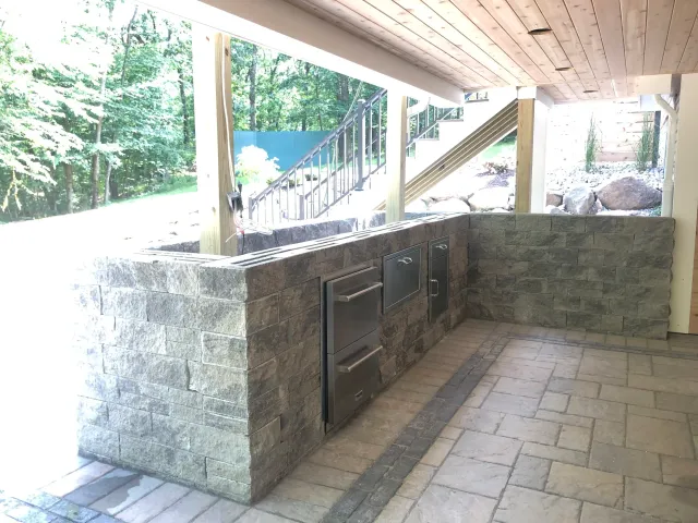Outdoor kitchen area with gray brick counter, built-in grill, and metal storage cabinets, on a stone tile patio, with a wooden staircase and landscaped yard visible in the background.