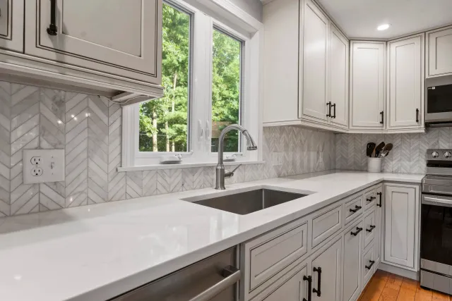White kitchen with marble backsplash, white cabinets, stainless steel sink, window showing green trees outside, and a stainless steel stove.