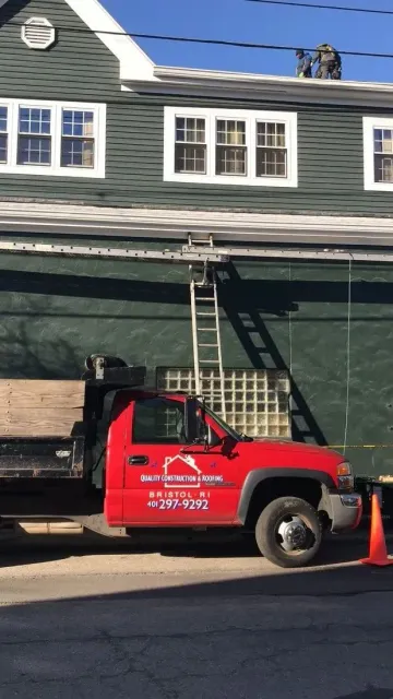 A red dump truck parked in front of a dark green building with a ladder leaning against its wall.
