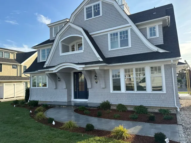 A large, newly constructed, multi-story house with gray shingles, white trim, and a black roof. The front yard has a curved pathway, small bushes, and a well-maintained lawn.