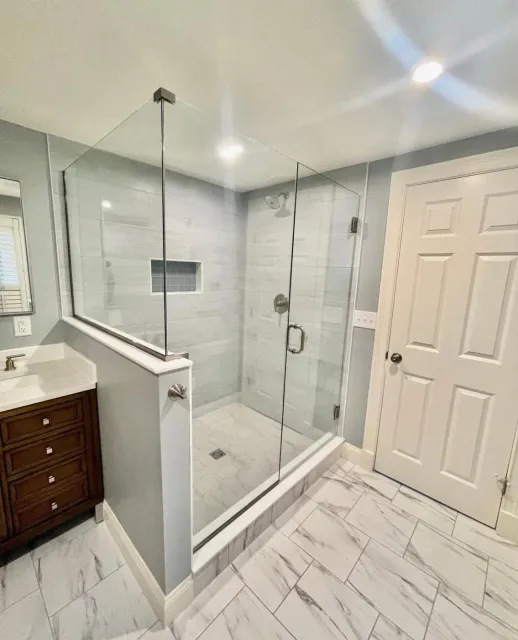 Modern bathroom with a glass-enclosed shower featuring light gray tiles and a built-in niche, a white marble floor, and a wooden vanity with a white countertop.