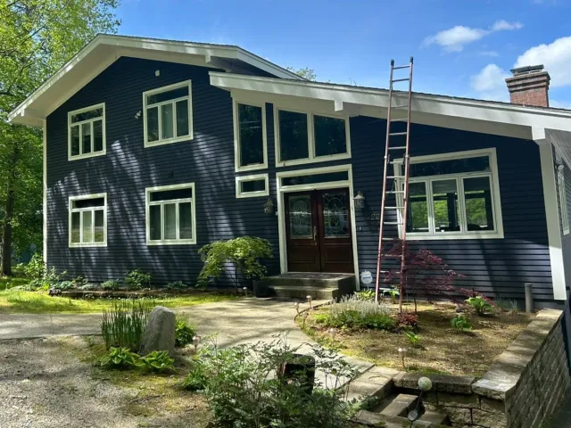 Dark blue house with white trim and front porch, viewed from a gravel driveway on a sunny day
