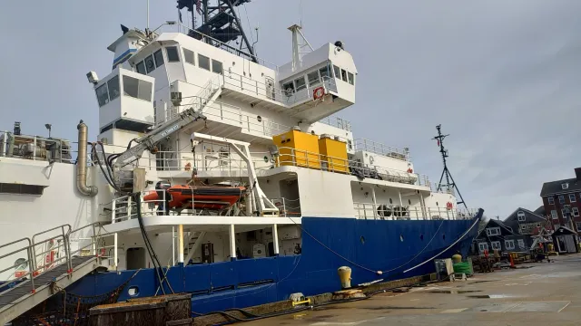 Large white and blue ship docked at a harbor with buildings and cloudy sky in the background.