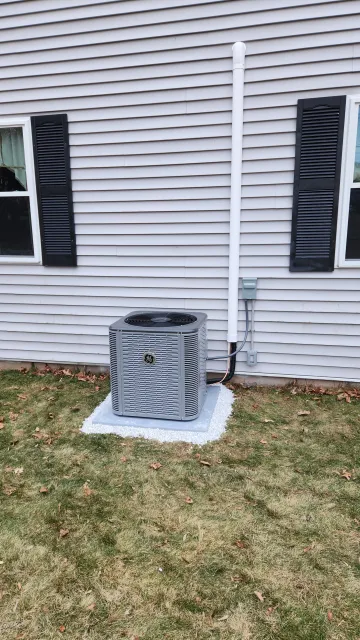 Exterior view of a house with white vinyl siding, two small windows with black shutters, and an outdoor HVAC unit on a concrete slab surrounded by white gravel, with a drain pipe running vertically along the wall.