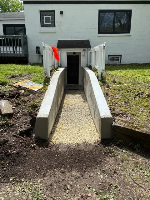 Concrete accessibility ramp leading to a basement entrance on a white house, with gravel path and red flagging tape