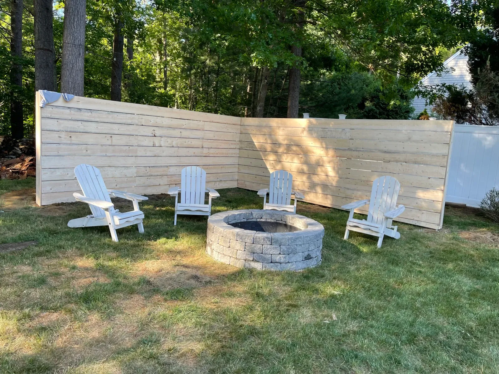 Outdoor backyard area with a wooden privacy fence, several white Adirondack chairs, a fire pit, lush green grass, and trees in the background.