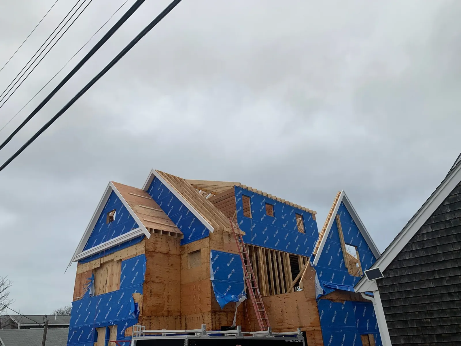 A house under construction with wooden framing and blue weatherproofing wrap, with roof trusses in place, in cloudy weather. Scituate, Norwell, Hanover, Hanson, Marshfield, South Shore. Massachusetts Local Construction Company.