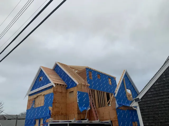 A house under construction with wooden framing and blue weatherproofing wrap, with roof trusses in place, in cloudy weather. Scituate, Norwell, Hanover, Hanson, Marshfield, South Shore. Massachusetts Local Construction Company.