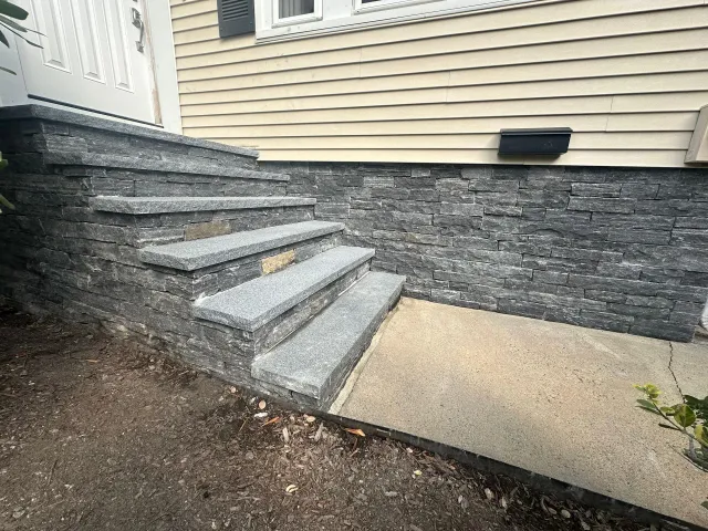 Concrete steps leading to a house entrance beside a stone retaining wall and beige siding