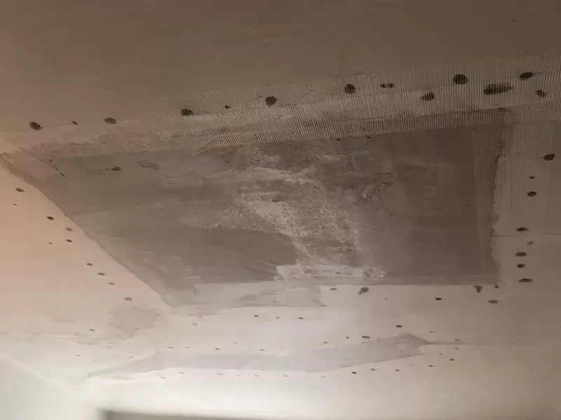 A ceiling with patches of different textures and colors, showing evidence of repair work with exposed drywall tape and numerous screw holes.