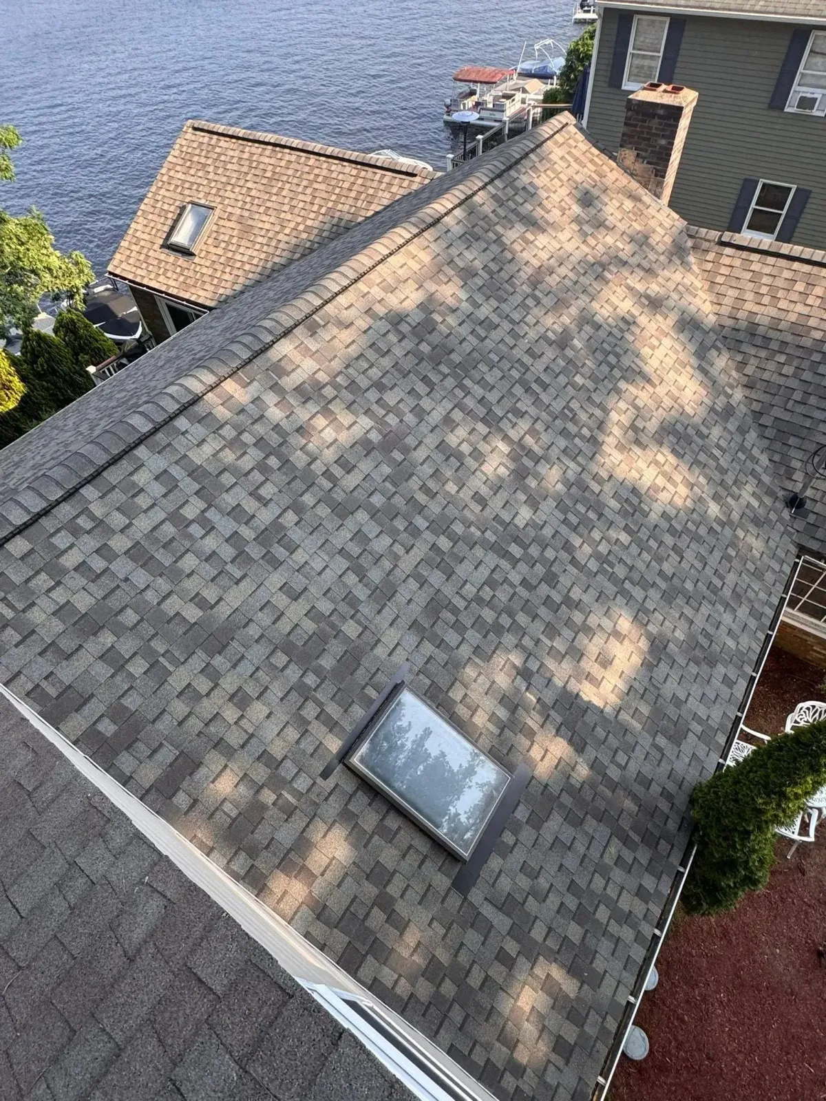 Aerial view of a gray shingle roof with a skylight, beside water and neighboring houses.