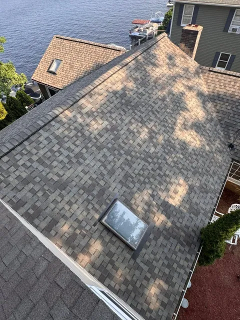 Aerial view of a gray shingle roof with a skylight, beside water and neighboring houses.