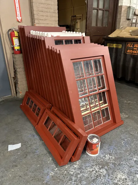 Red-painted wooden window frames leaning against a wall in an industrial or storage area, with a fire extinguisher and a large trash bin nearby.