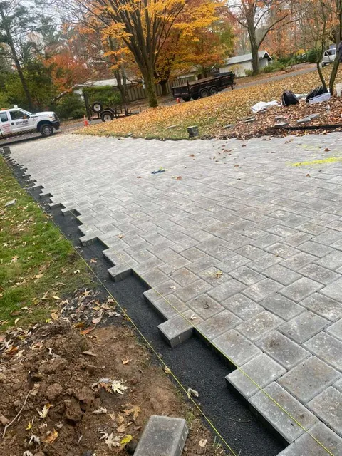A partially installed gray paver driveway under construction with a black border, set against an autumn-colored landscape.