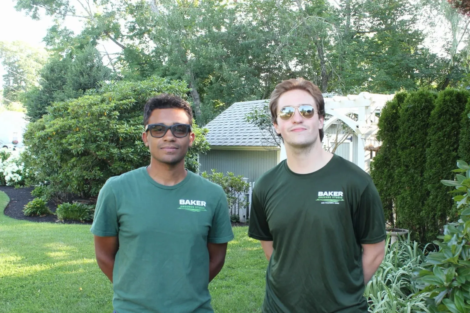 Two young men standing outdoors in a backyard with green trees and bushes behind them. They are wearing matching dark green T-shirts with white text that reads 'Baker Grounds Studio.' The man on the left has short dark hair, glasses, and a slight smile. The man on the right has medium-length brown hair, sunglasses, and a slight smile.