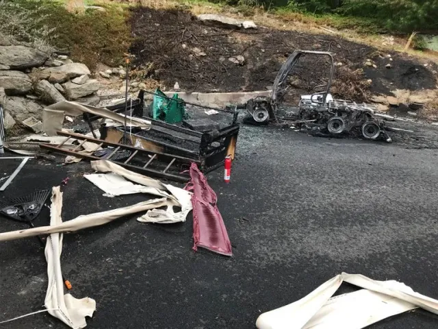 Charred remains of a vehicle and debris scattered on asphalt, with burnt hillside in the background.