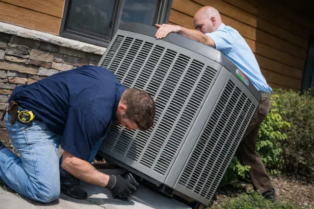 Two technicians installing an outdoor air conditioning unit next to a stone-walled building.