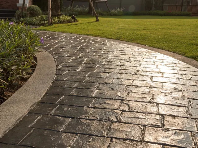Wet brick pathway curving through a well-manicured garden with green grass and plants, in sunlight.