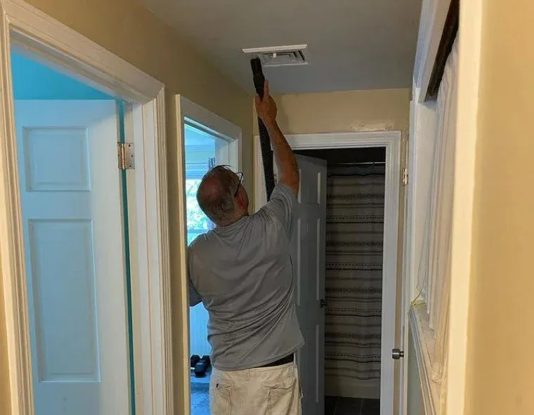Man cleaning or inspecting a ceiling vent in a hallway with a duster.