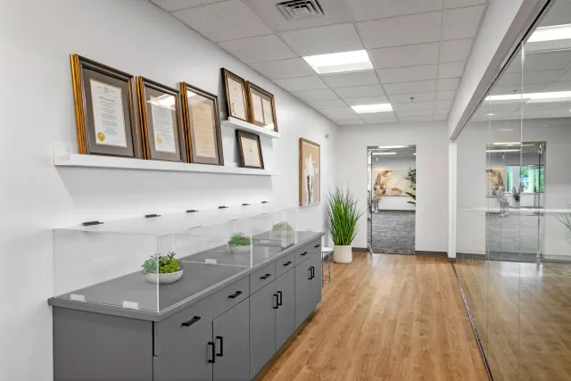 Office hallway with framed certificates on the wall, a gray cabinet with plants inside a glass display case, and a potted plant near the door.