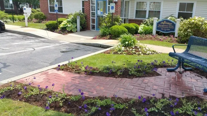 A blue bench sits next to a brick walkway surrounded by purple flowers and greenery in front of an apartment building.